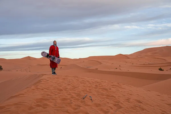 Man in traditional clothes with sandboard standing on sand dunes against sky — Zdjęcie stockowe