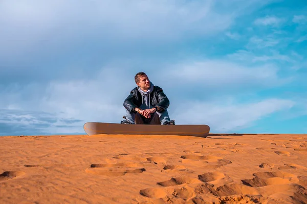 Man with sandboard sitting on sand in desert against cloudy sky — Foto Stock