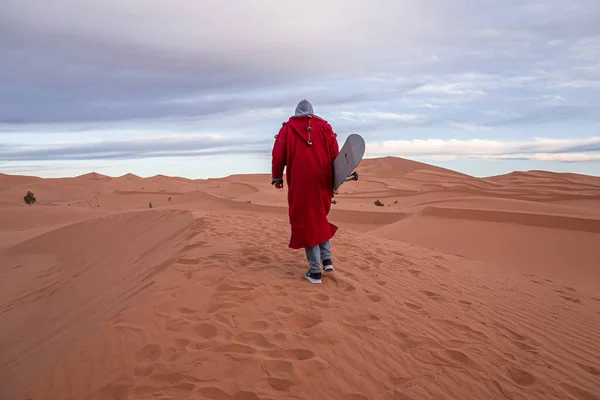 Man in traditional clothes with sandboard walking on sand dunes against sky — Foto Stock