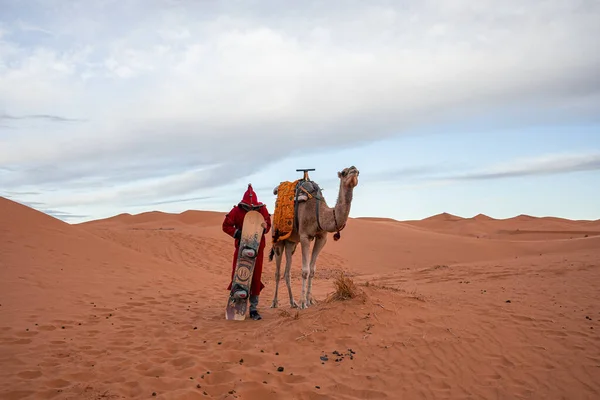 Man in traditional clothes with sandboard standing beside camel in desert — Stockfoto