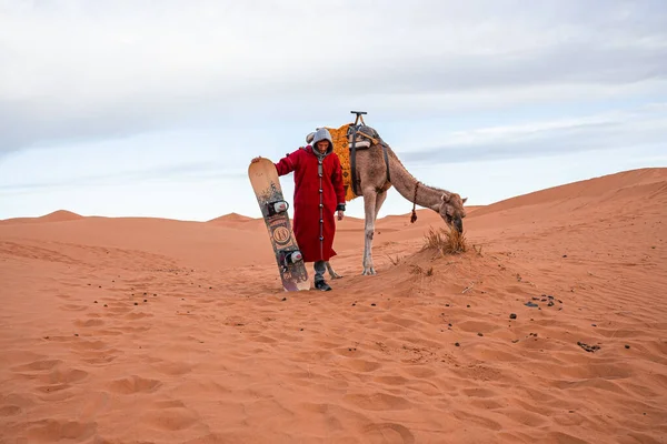 Man in traditional clothes with sandboard standing beside camel in desert — Stok fotoğraf