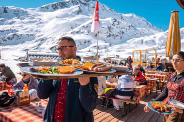Aziz Anton am Arlberg. 10 Mart 2022. Garsonlar açık kafede masaların yanında yürürken tepsileri taze yiyecekle birlikte taşıyorlar, garsonlar güneşli bir günde dağ kafede tabakları taşıyorlar.