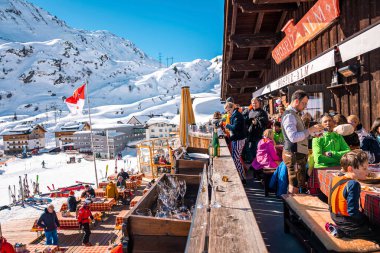 Aziz Anton am Arlberg. 10 Mart 2022. Kayak tatili sırasında kayak merkezindeki masaların yanındaki açık kafede oturan insanlar güneşli bir günde dağ kafesinde oturan turistler.