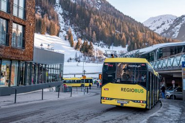 Aziz Anton am Arlberg. 10 Mart 2022. Kayak tatili sırasında ana caddede kayak otobüsü, modern otelin yanındaki yolda karlı dağlara karşı sarı otobüs.