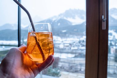 Kadehte köpüklü bir içecek tutarken yakın plan. Erkek turistler lüks otellerde soğuk içeceklerin tadını çıkarıyorlar. Arka plandaki cam pencereden görülen dağların görüntüsü.