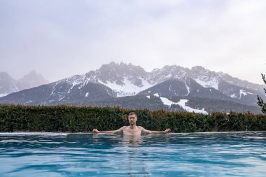 Adam kış boyunca sıcak havuzda yüzüyor. Lüks kayak merkezindeki jakuzide dinlenen erkek turist. Gökyüzüne karşı karla kaplı dağlar.