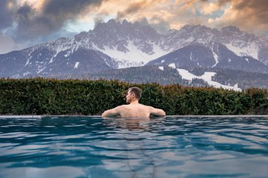 Adam dağ manzaralı havuzda eğleniyor. Lüks oteldeki jakuzide yüzen turistlerin arka manzarası. Gün batımında gökyüzüne karşı karla kaplı manzara.