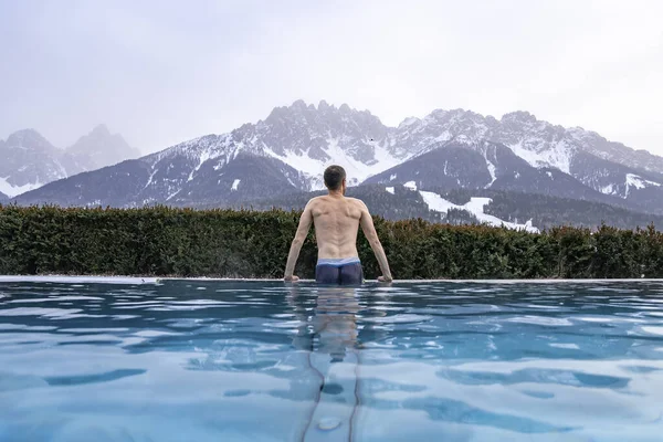 Dağların manzaralı sıcak havuzda yüzen bir turist. Lüks bir tatil köyünde suyun tadını çıkaran bir adamın arka görüntüsü. Kar kış boyunca gökyüzünü kapladı.