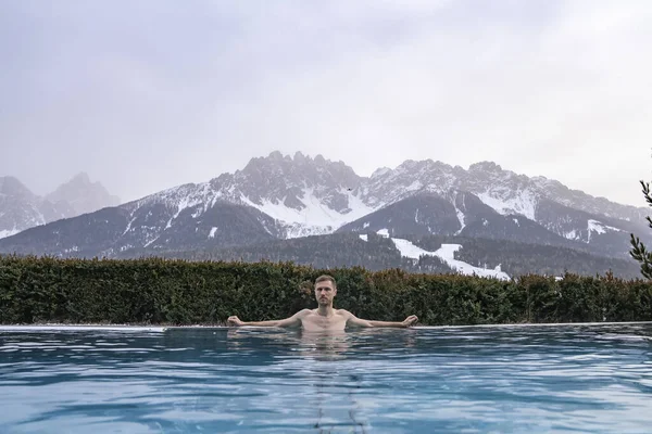 Adam kış boyunca sıcak havuzda yüzüyor. Lüks kayak merkezindeki jakuzide dinlenen erkek turist. Gökyüzüne karşı karla kaplı dağlar.
