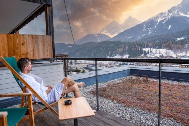 Tourist looking at sunrise over snowcapped mountains. Man wearing bathrobe is sitting on chair. He is relaxing at luxurious hotel during winter. clipart