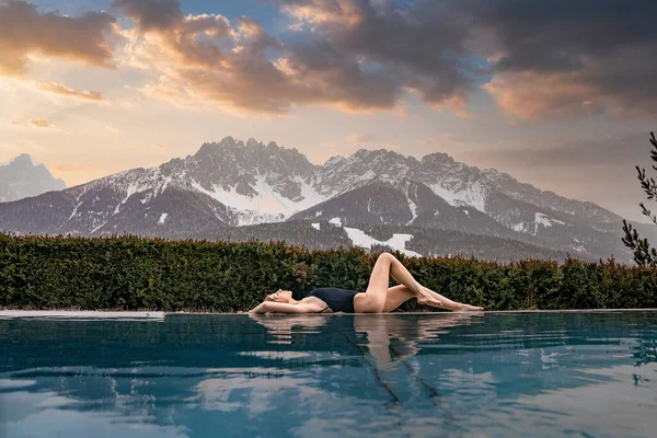 Havuz kenarında mayo giymiş bir kadın yatıyor. Gün batımında gökyüzüne karşı karla kaplı dağlar. Bayan turist kış boyunca lüks bir otelde tatilin tadını çıkarıyor..