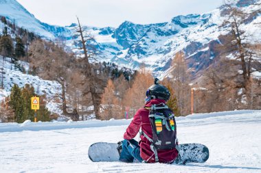 Kar üzerinde dinlenen yorgun bir snowboardcu. Güneşli bir günde gökyüzüne karşı muhteşem dağ manzarası. Kış boyunca karda oturan turist.