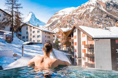 Matterhorn dağlarına ve evlerine bakarken havuzda yüzen bir turist. Jakuzide rahatlayan bir adamın arka görüntüsü. Kış boyunca Alp Dağları 'nda kar manzarası.