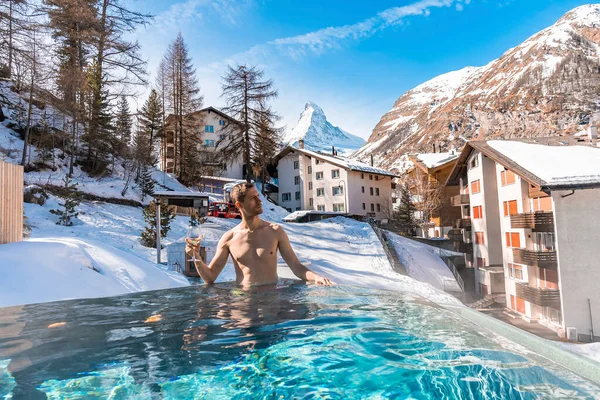 Havuzda yüzerken şarap içen bir adam. Matterhorn Dağı 'na ve evlere karşı jakuzide dinlenen turistler. Alp bölgesindeki karlı manzaranın manzarası.