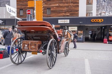 Zermatt, İsviçre. 15 Mart 2022. Sokakta bagajları olan turistler bekliyor. Dükkanların önünde geleneksel at yarışı. Alp bölgesindeki kasaba meydanında hayvan çizilmiş araç.