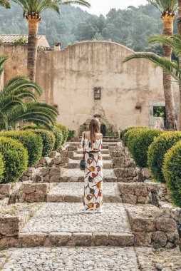 Çiçeklerin arasında basamaklarda duran dişi turistler. Günlük elbiseli bir kadın tarihi parkı keşfediyor. Yaz tatilini adada geçiriyor..