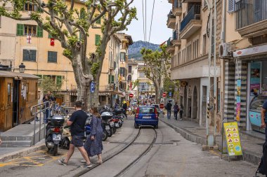 Mallorca, İspanya. 27 Nisan 2022. Turistler, binaların arasındaki rayları geçerken konuşuyor. Araba şehirde demiryolunda hareket ediyor. Eski şehrin dar sokaklarında yürüyen insanlar.