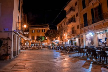 Mallorca, İspanya. 27 Nisan 2022. Kasaba meydanındaki açık hava restoranları ve kafeler aydınlatılmış. Şehirde gökyüzüne karşı kurulmuş bir otel binası. Gece boyunca şehir manzarası.