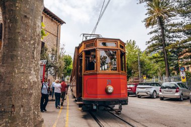 Mallorca, İspanya. 27 Nisan 2022. Geleneksel tramvay tren raylarında hareket ediyor. Erkek ve kadın turistler tarihi turuncu trenin yanından geçiyorlar. Şehre park etmiş arabalar gökyüzüne karşı.