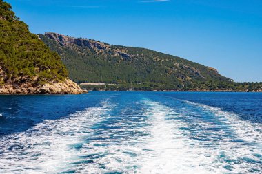 Surf created by wake in sea. Beautiful view of cliff at island against blue sky. Water moving in Mediterranean ocean during sunny day.