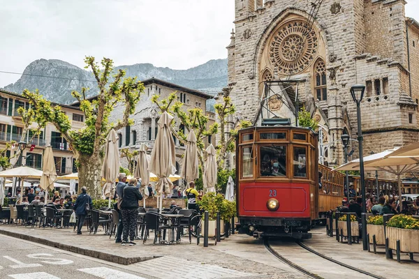 Mallorca, İspanya. 27 Nisan 2022. Tramvay ortaçağ kilisesine karşı tren raylarında ilerliyor. Erkek ve kadın turistler kentteki restoranları keşfediyorlar. Sant Bartomeu de Soller tarihi şehirdedir..
