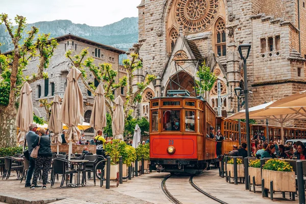 Turuncu tramvay kilise ve restoranlar arasında tren raylarında hareket ediyor. Şehri keşfeden erkek ve kadın turistler. Tarihi şehirde Sant Bartomeu de Soller Kilisesi.