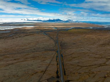 Güneşli hava şartlarında İzlanda 'nın bulutlu dağlarına ve tepelerine uzanan sonsuz yol. Havadan sinematik görünüm.