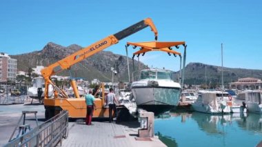 Sarı vinç limanda yelkenliyi kaldırıyor. İşçiler ağır makineler kullanarak denizden gemiler alıyorlar. Tersanedeki portatif asansör temiz gökyüzüne karşı..