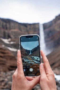 Hengifoss 'u fotoğraflayan turist görüntüsü. Akıllı telefon ekranında dağların ortasındaki güzel şelale. Gezgin tatilde volkanik vadiyi keşfediyor..