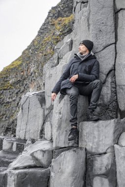 Turistler bazalt sütunlarda otururken gözlerini kaçırıyorlar. Deniz kıyısında kayalık oluşumda rahatlayan bir adam. Ünlü Reynisfjara Sahili 'ndeki gri desenli taşların manzarası..
