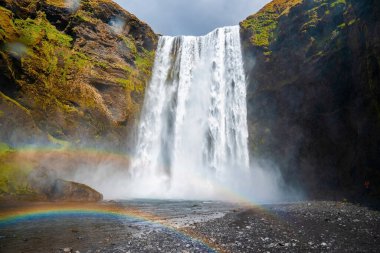 Skogafoss şelalesinin güçlü çağlayanlarına karşı alçak açılı güzel gökkuşağı manzarası. Volkanik vadideki dağdan akan nehir. Bulutlu gökyüzüne karşı manzaranın doğal vahşi doğası.
