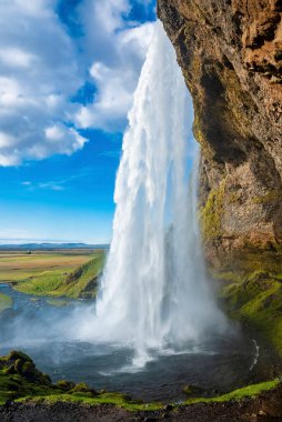 Seljalandsfoss 'un dağdan akan güzel çağlayanları. Mavi bulutlu gökyüzüne karşı muhteşem bir şelale. Vadideki ünlü turistlerin manzarası.