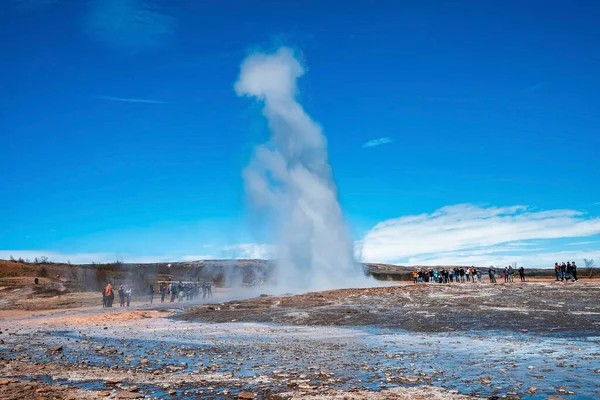 Strokkur gayzer patlamasının tadını çıkaran turistler. Mavi gökyüzüne karşı dramatik manzaradan yayılan duman. Erkekler ve kadınlar güneşli bir günde vadideki ünlü eğlenceyi keşfediyorlar..