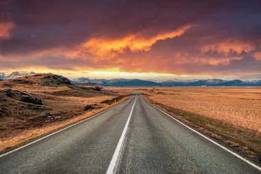 Volkanik manzaranın ortasında kaybolan boş caddenin görüntüsü. Dramatik gökyüzüne karşı sıradağların manzarası. Gün batımında azalan yolda yol işaretleri.