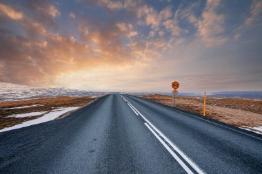 Karla kaplı arazide yol azalıyor. Gün batımında boş caddede yol işaretleri. Yol kenarında bulutlu gökyüzüne karşı tabela olan otobanın manzarası.