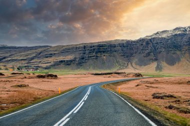 Dağ sırasına doğru giden boş yol manzarası. Gün batımında volkanik manzaranın ortasında caddedeki yol işaretleri. Bulutlu gökyüzüne karşı vadide azalan otoyolun manzarası.