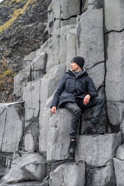 Güzel bazalt sütunlarda oturan düşünceli bir turist. Kayalık formasyonda rahatlarken gözlerini kaçıran bir adam. Ünlü Reynisfjara sahilindeki gri desenli taşların manzarası..