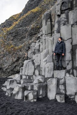 Uçurumlardaki bazalt sütunlarda duran gezgin. Siyah deniz kıyısında kaya oluşumunu keşfeden erkek turist. Ünlü Reynisfjara Sahili 'ndeki desenli taşların manzarası..