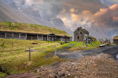 Görkemli Vestrahorn dağının altındaki çimenli tarlaların ortasındaki geleneksel evler. Stokksnes yakınlarında bulutlu gökyüzüne karşı Viking köyü manzarası. Günbatımında volkanik vadide tarihi turistik cazibe.