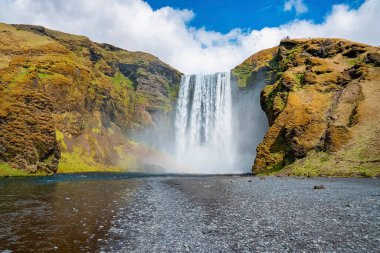 Vadideki Skogafoss şelalesinin güçlü şelaleleri. Uçurumların arasından akan güzel nehir suyu mavi gökyüzüne karşı. Volkanik manzaranın ideal doğal manzarası.