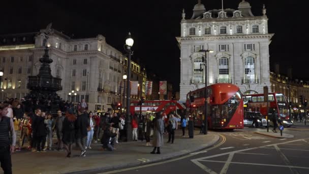 ロンドンのピカデリー サーカスでの夜の生活 通りを横断する人々とピカデリーサーカスを通って運転する広告バナーと象徴的な赤いダブルデッキバス — ストック動画