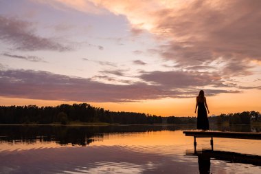 Lonely girl silhouette at sunset by the lake. Dreamy lady emjoying orange sunset by the lake in a pier. Girl enjoying magical nature.