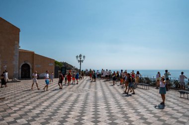 Taormina, Sicilya, İtalya. 26 Ağustos 2022. Erkek ve kadın turistler güneşli bir günde eski kasaba meydanında eğleniyorlar. Kıyıdaki tarihi binaların arkasında mavi gökyüzü var..