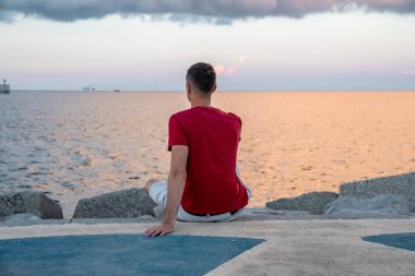 Erkek turistin gezinti güvertesinde dinlenmesi. Genç adam deniz kenarında otururken sahile bakıyor. Tatilde boş zamanlarını geçirirken gündelik giysiler giyiyor..