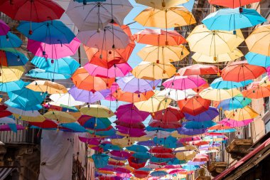 Catalina, Sicilya, İtalya. 26 Ağustos 2022. Arka planda gökyüzü olan binaların arasında asılı çok renkli şemsiyeler. Yaz boyunca yerleşim bölgesindeki caddelerde dekoratif sanat manzarası.