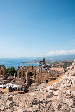 Akdeniz kıyısındaki antik Yunan tiyatrosunun kalıntılarındaki kayalar. Giardini Naxos körfezinde, arkasında mavi gökyüzü olan bir bina. Güneşli bir günde tarihi turistik mekan manzarası.