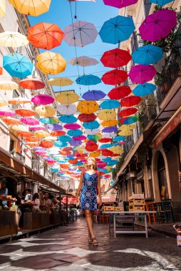 Catalina, Sicilya, İtalya. 26 Ağustos 2022. Caddenin karşısındaki çok renkli şemsiyelere bakan kadın turist. Yaz boyunca şehirde güzel dekorasyonların keyfini çıkarırken bir kadın kafenin önünden geçiyor..