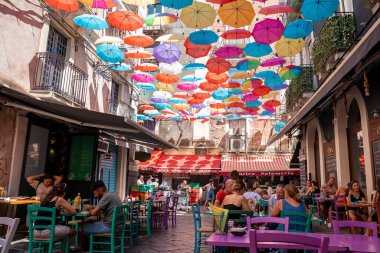 Catalina, Sicilya, İtalya. 26 Ağustos 2022. Erkek ve kadın turistler kafelerde renkli şemsiyelerin altında binaların arasında dinleniyorlar. Yaz boyunca şehrin sokaklarındaki güzel dekorasyon manzarası.