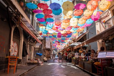 Catalina, Sicilya, İtalya. 26 Ağustos 2022. Binaların arasında caddelerin üzerinde çok renkli şemsiyeler asılı. Erkek ve kadın turistler yaz boyunca şehirde güzel dekorasyonlar altında kafede dinleniyorlar..