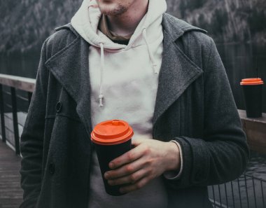 Paltolu bir adam, ormanın ve nehrin bulanık arka planında bir fincan kahve tutuyor.